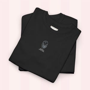 Puede incluir: Dos camisetas negras apiladas, con la camiseta superior doblada para revelar un pequeño gráfico blanco de una máscara y el texto "DAMON'S LITTLE DEVIL". Las camisetas están sobre un fondo rosa claro.