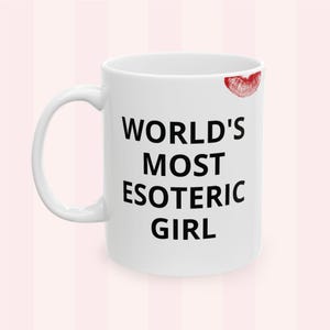 Può includere: Tazza in ceramica bianca con la scritta "WORLD'S MOST ESOTERIC GIRL" in nero. Un segno di bacio di rossetto rosso si trova in alto a destra della tazza. La tazza ha un manico curvo.