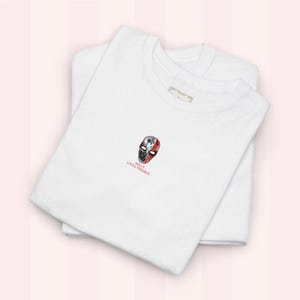 May include: Two folded white t-shirts. The top shirt has a small embroidered design of a split mask in black, red, and white, with the text "WILL'S LITTLE TROUBLE" beneath. The shirts are stacked on a pink background.