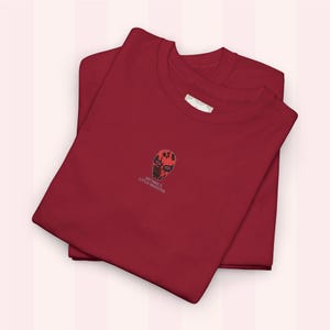 Könnte beinhalten: Zwei weinrote T-Shirts sind ordentlich gefaltet, eines auf dem anderen. Das obere Shirt hat ein kleines gesticktes Design einer rot-schwarzen Totenkopfmaske mit dem Text "MICHAEL'S LITTLE MONSTER" darunter. Die Shirts scheinen aus weichem Material zu sein.