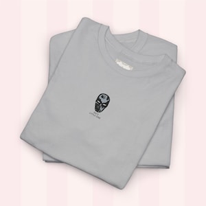 Könnte beinhalten: Zwei hellgraue T-Shirts sind ordentlich gefaltet und gestapelt. Jedes Shirt hat ein kleines, gesticktes schwarz-graues Maskendesign mit dem Text "KAY'S LITTLE ONE" darunter. Die Shirts befinden sich auf einem rosa Hintergrund.
