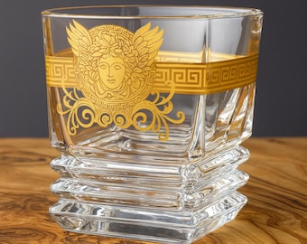 May include: A clear, square-shaped glass with a gold band featuring a Medusa head design. The glass has a stacked, textured base and is suitable for serving beverages. The gold accents provide a luxurious look.