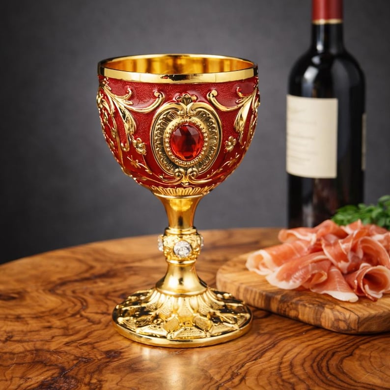 May include: Ornate, gold-colored goblet with a ruby-red bowl and a large, central red gem. The goblet has intricate gold detailing and a decorative base. A bottle of red wine and a wooden cutting board with sliced meat are in the background.