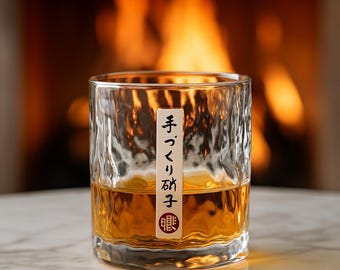 May include: A clear, textured glass filled with amber liquid, possibly whiskey or a similar spirit. A white label with Japanese text is affixed to the glass. The background features a blurred fireplace with warm orange and yellow flames.