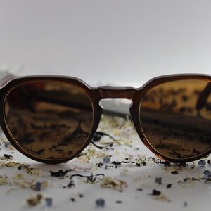 Puede incluir: Gafas de sol con montura marrón y lentes redondas. Las gafas tienen un puente de cerradura y lentes tintadas. El fondo presenta elementos florales dispersos.