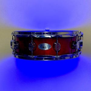 Pode incluir: Caixa vermelha com ferragens cromadas. O tambor tem um acabamento polido e apresenta o texto "cadp M5" numa pequena placa de metal. O tambor é iluminado com luz azul.