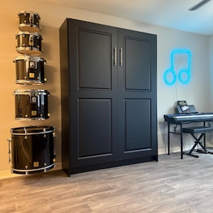 May include: A black drum set with silver hardware is mounted on a wall next to a large black cabinet. A neon blue musical note is on the wall. A keyboard and bench are in the background.