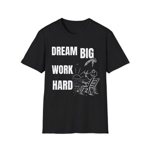 Puede incluir: Camiseta negra con el texto blanco "DREAM BIG WORK HARD". Un muñeco de palo se relaja en una playa, sosteniendo una bebida, con una puesta de sol de fondo. La camiseta está hecha de un material suave.
