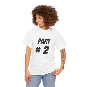 Puede incluir: Camiseta blanca con el texto "PART # 2" en letras negras en negrita. La camiseta es de estilo cuello redondo clásico y parece estar hecha de un material suave y cómodo. El texto está centrado en la parte delantera de la camiseta.