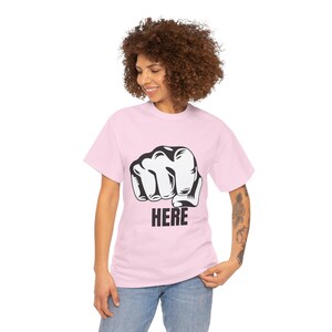 Puede incluir: Camiseta rosa claro con un gráfico de puño cerrado en blanco y negro y la palabra "HERE" impresa debajo. La camiseta está hecha de un material suave.
