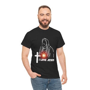 Puede incluir: Camiseta negra con un contorno blanco de una figura religiosa, una cruz y el texto "I LOVE JESUS". El diseño tiene un efecto de brillo rojo. La camiseta es de cuello redondo.
