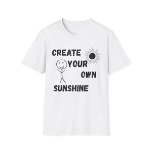 Puede incluir: Camiseta blanca con la frase negra "CREATE YOUR OWN SUNSHINE". El diseño incluye un muñeco de palo con una carita sonriente y un sol. Un diseño sencillo y motivador.