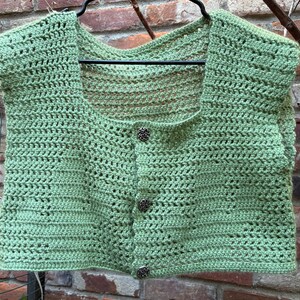 May include: A light green, hand-crocheted vest with a square neckline and cap sleeves. The vest features a button-up front with four decorative buttons. The open weave design and soft green color give it a summery, handmade feel.