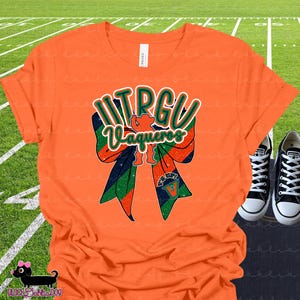 May include: An orange t-shirt featuring a large graphic of a glittery bow in orange, green, and dark blue. The text UTRGU Vaqueros is printed in a stylised font across the bow, with a small mascot silhouette and the text V's UP visible below.