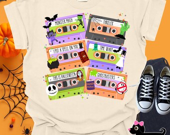 Puede incluir: Camiseta color crema con un collage de cintas de cassette con diseños y texto de Halloween. Las cintas son de varios colores, incluyendo morado, naranja y verde, con frases como "Monster Mash" y "Thriller". Se ven zapatillas Converse negras.