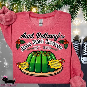 May include: Red sweatshirt with the text "Aunt Bethany's Jello Mold Company EST. 1989". The graphic includes a green jello mold, holly, and the phrases "Extra Crunchy" and "I don't know about the cat, but I sure am enjoying it - Cousin Eddie".