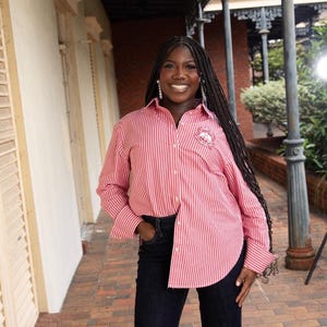 May include: A woman wearing a red and white striped button-down shirt with a small embroidered design on the chest pocket. She is also wearing dark blue jeans. The background includes a brick walkway and a building with white walls.