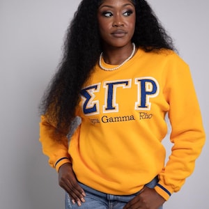 May include: Mustard yellow sweatshirt with blue and white Greek letters and the words "Sigma Gamma Rho". The sweatshirt has blue stripes on the cuffs. The person is wearing blue jeans and a blue headband.