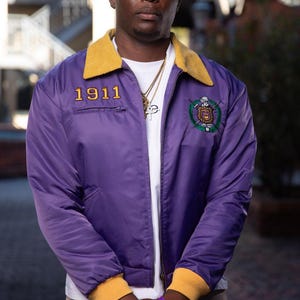May include: A purple jacket with a yellow collar and cuffs. The jacket has the number "1911" embroidered in gold on the chest and a crest on the left side. The person is wearing a white shirt and gold chain.
