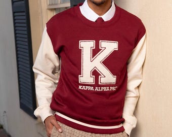 May include: A maroon and cream letterman-style sweatshirt with the Greek letter "K" and "KAPPA ALPHA PSI" in white. The person is wearing a white collared shirt and plaid pants.