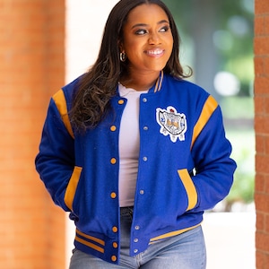 May include: A blue and gold varsity jacket with a crest patch. The jacket has gold stripes on the sleeves and a button-up front. The person is wearing a white shirt and denim shorts.