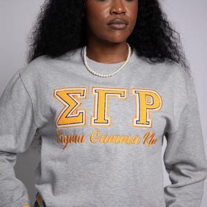 May include: A gray sweatshirt with the letters "ΣΓΡ" in yellow and orange embroidery, with the words "Sigma Gamma Rho" below. The sweatshirt has blue and yellow striped cuffs.