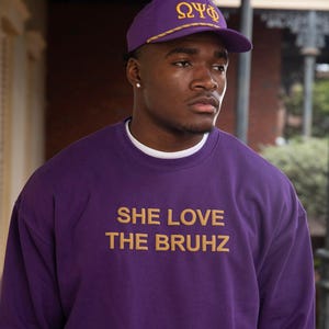 May include: A purple sweatshirt and cap with gold lettering. The cap displays Greek letters. The sweatshirt has the text "SHE LOVE THE BRUHZ" in gold. The person is wearing a white undershirt.