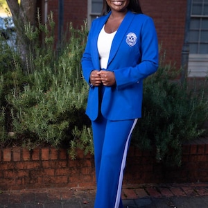 May include: A woman wearing a royal blue suit with a white top. The suit jacket has a crest on the chest. The pants have a white stripe down the side. The background includes a brick building and green foliage.