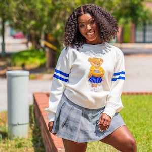 May include: Cream-colored sweatshirt with blue stripes on the sleeves and a teddy bear graphic. The bear wears a yellow shirt and blue skirt with the number 1922. Paired with a gray plaid pleated skirt.