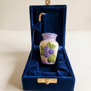 May include: A small, decorative urn with a lavender top and a cream-colored body, adorned with purple rose illustrations and green leaves. The urn is presented in a deep blue velvet box with a gold clasp and hinge.