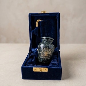 May include: A small, decorative urn with a tree of life design, presented in a deep blue velvet box. The urn is dark green with gold accents. The box has a gold clasp and a matching velvet interior.
