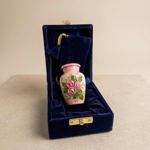 May include: A small, decorative urn with a pink rim and floral design, presented in a dark blue velvet box. The urn features pink roses and green leaves. The box has a gold clasp and a matching velvet interior.