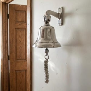 May include: A polished silver bell mounted on a white wall. The bell is attached to a bracket and has a braided rope pull. The bell is near a wooden door frame.