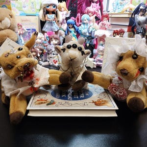 May include: Three plush animal figures with multiple eyes, arranged on a book. The figures are brown and tan, with white collars and decorative accents. A variety of dolls and figurines are visible in the background.