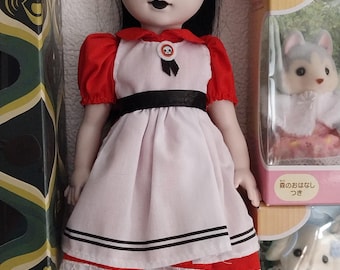 Puede incluir: Muñeca con rostro pálido, cabello negro y diadema roja. La muñeca viste un vestido rojo y blanco con una cinta negra y zapatos negros. La muñeca tiene una expresión espeluznante con maquillaje oscuro y una marca de belleza.