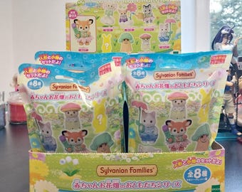 May include: A display box of Sylvanian Families collectible figures. The box features colorful packaging with images of various animal characters in floral themes. The text on the box is in Japanese. The box contains multiple blind bags.