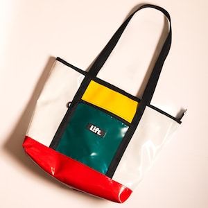 Può includere: Una borsa tote bianca con un design a blocchi di colore. La borsa presenta una base rossa, una tasca verde e gialla e un manico nero. La parola "Lift." è stampata sulla tasca verde.