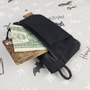 Puede incluir: Pequeño estuche negro con cremallera, un pequeño lazo y una solapa rectangular. El estuche está abierto, revelando un billete de dólar y una tarjeta de crédito dorada. Fondo gris claro con ilustraciones de Batman.