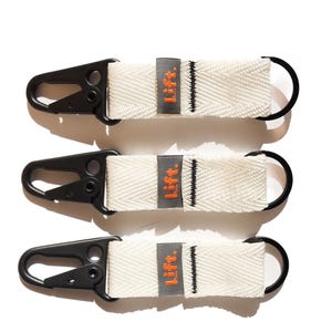 Puede incluir: Tres llaveros blancos con mosquetones negros y anillos negros. Cada llavero tiene una etiqueta gris con la palabra "Lift." en naranja. Los llaveros están hechos de un material tejido.