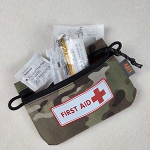 Puede incluir: Un estuche de primeros auxilios con patrón de camuflaje, cremallera negra y una etiqueta blanca con el texto rojo FIRST AID y una cruz roja. El estuche está abierto y muestra suministros médicos como una toallita con alcohol.