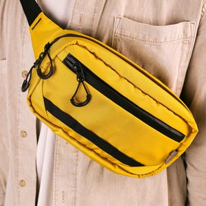 Puede incluir: Un bolso de cintura amarillo con cremalleras y detalles en negro. El bolso tiene forma rectangular con la parte superior curvada y una correa negra. El bolso de cintura está hecho de un material texturizado y se lleva sobre el pecho.