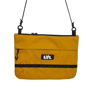May include: Mustard yellow crossbody bag with black accents. Features a black zipper, a black strap, and the word "Lift." in a white rectangle. The bag is rectangular and has a black strap at the bottom.