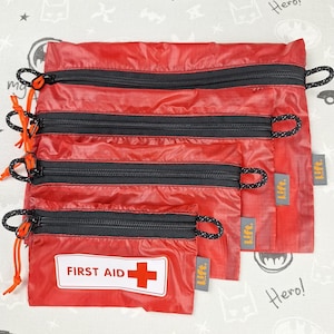 May include: A set of four red nylon zippered pouches. The smallest pouch has a white rectangle with "FIRST AID" and a red cross. Each pouch has a black zipper and a black and orange pull cord.