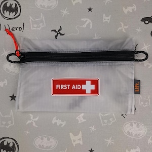 May include: A small, translucent grey first aid kit with a black zip and red pull cord. A red and white "FIRST AID" label is on the front. The kit is on a grey fabric with Batman-themed patterns.