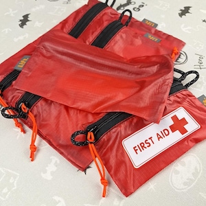 May include: A collection of red nylon pouches with black zips and orange cords. One pouch features a white label with "FIRST AID" and a red cross. The pouches are displayed on a patterned surface with the word "Hero!" visible.