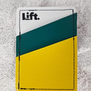 May include: A rectangular card holder with a white top section, a diagonal green stripe, and a yellow bottom. The word "Lift." is printed in black at the top. The edges are stitched with black thread.