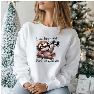 May include: White sweatshirt featuring a cartoon sloth holding an "OUT OF ORDER" sign. The sloth is brown and tan, with the text "I am temporarily" and "Please try again later" printed above and below the sloth illustration.
