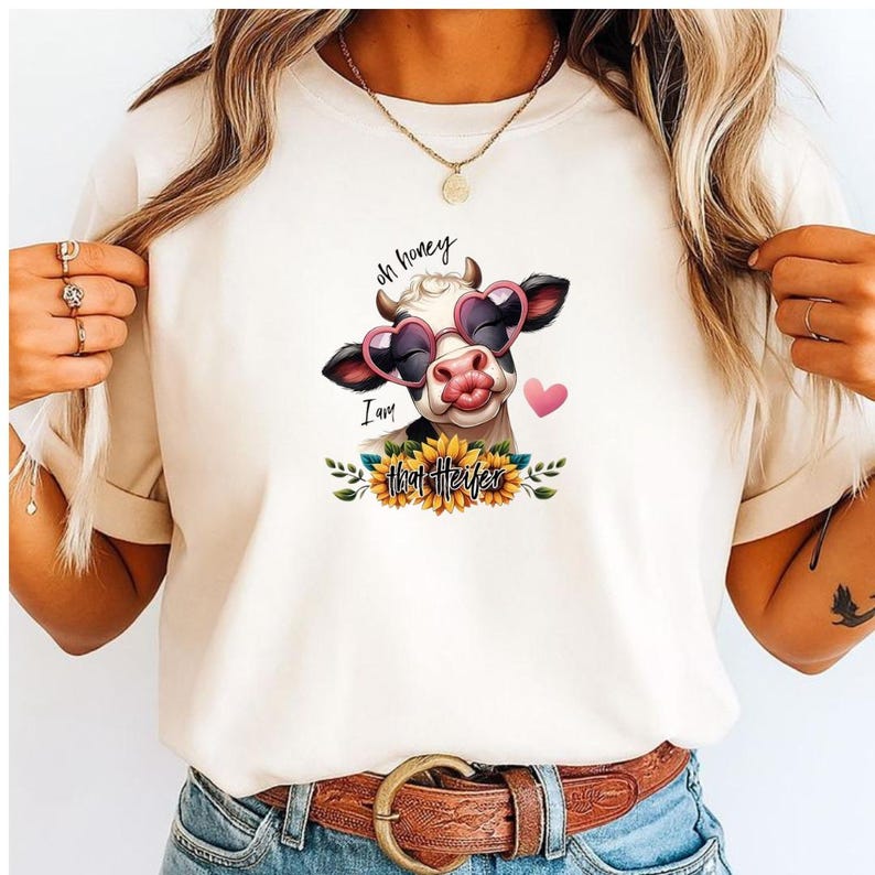 May include: Cream-colored t-shirt featuring a graphic of a cow wearing heart-shaped sunglasses and a kiss. The text "oh honey, I am that Heifer" is above and below the cow. Sunflowers and leaves surround the text.