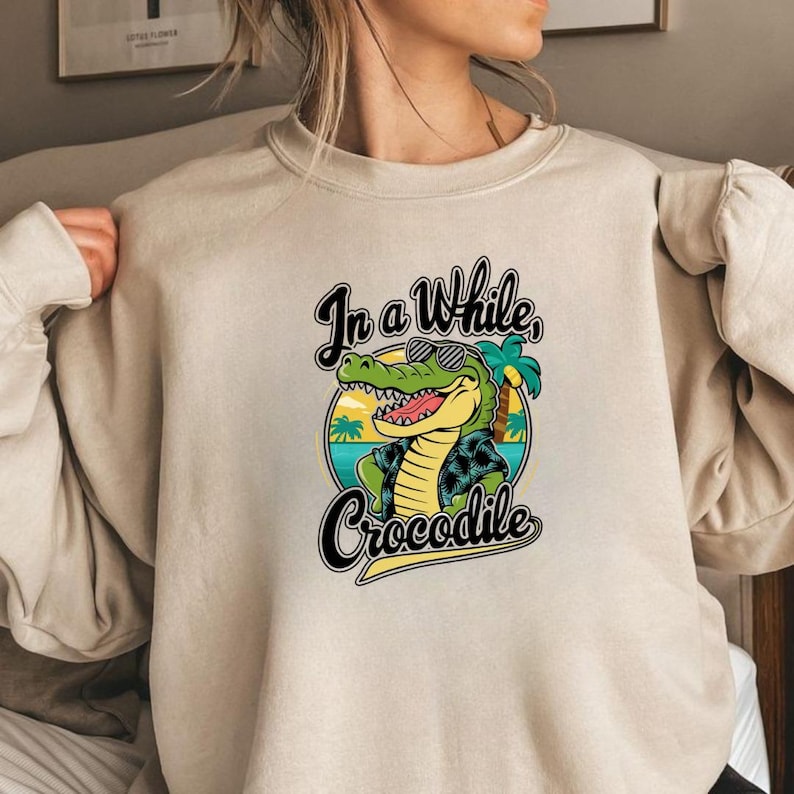 May include: Beige sweatshirt featuring a cartoon crocodile wearing sunglasses and a Hawaiian shirt. The graphic includes the text "In a While, Crocodile" in a stylized font, with a tropical beach scene in the background.