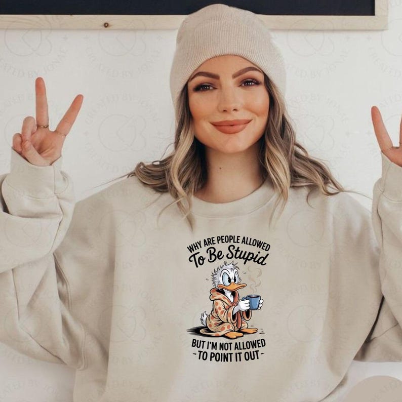 May include: Beige sweatshirt featuring a graphic of Donald Duck holding a mug, with the text "WHY ARE PEOPLE ALLOWED TO BE STUPID BUT I'M NOT ALLOWED - TO POINT IT OUT-". The sweatshirt is paired with a beige beanie.